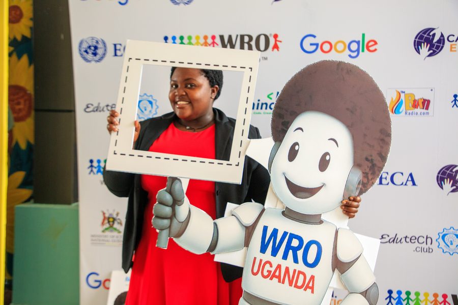 A participant holding a photo frame next to the WRO Uganda mascot at the Uganda National Championship